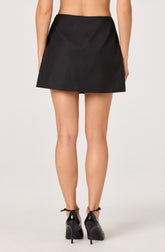 The image shows a woman wearing a black mini skirt that is high-waisted with a smooth finish, without visible pockets or embellishments. She is also wearing black high-heeled shoes with ankle straps. The background is plain and light-colored.